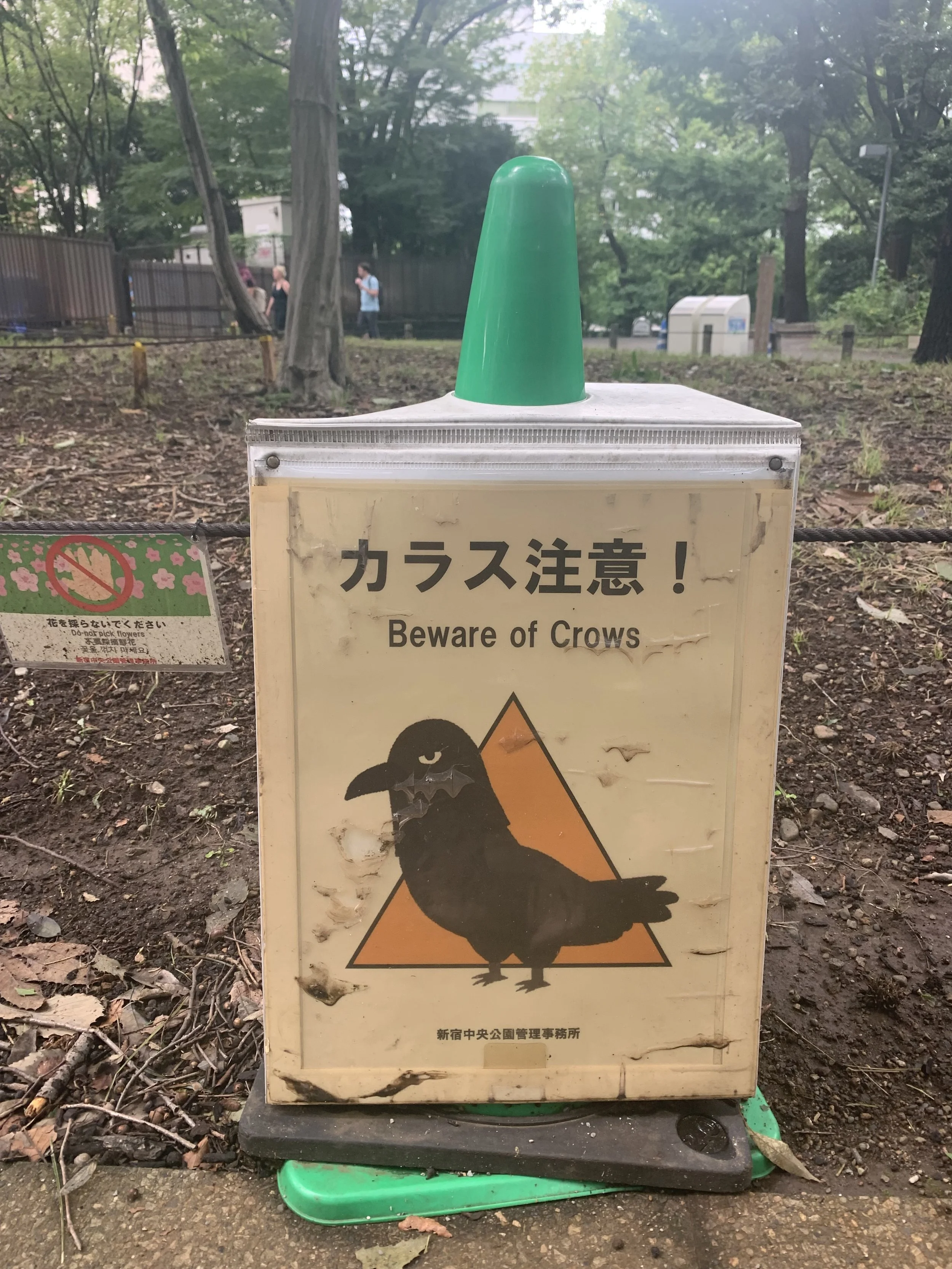 A sign warning visitors to beware of crows, featuring an illustration of a crow with a grim expression and a caution triangle in the background, placed outside in a park area.