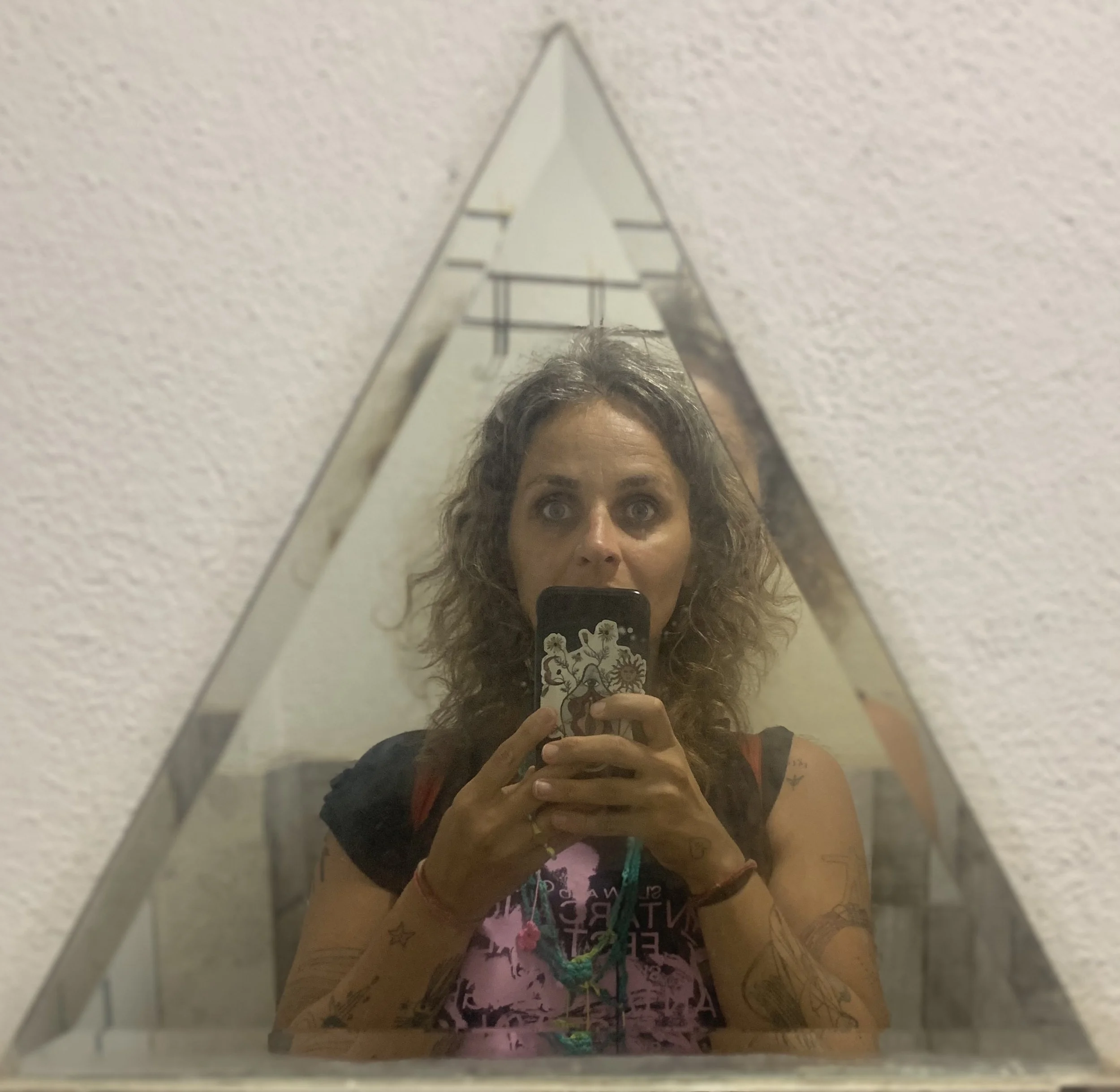 Woman with curly hair taking a mirror selfie in a triangular mirror, wearing a black T-shirt with pink and blue graphics, and multiple necklaces.