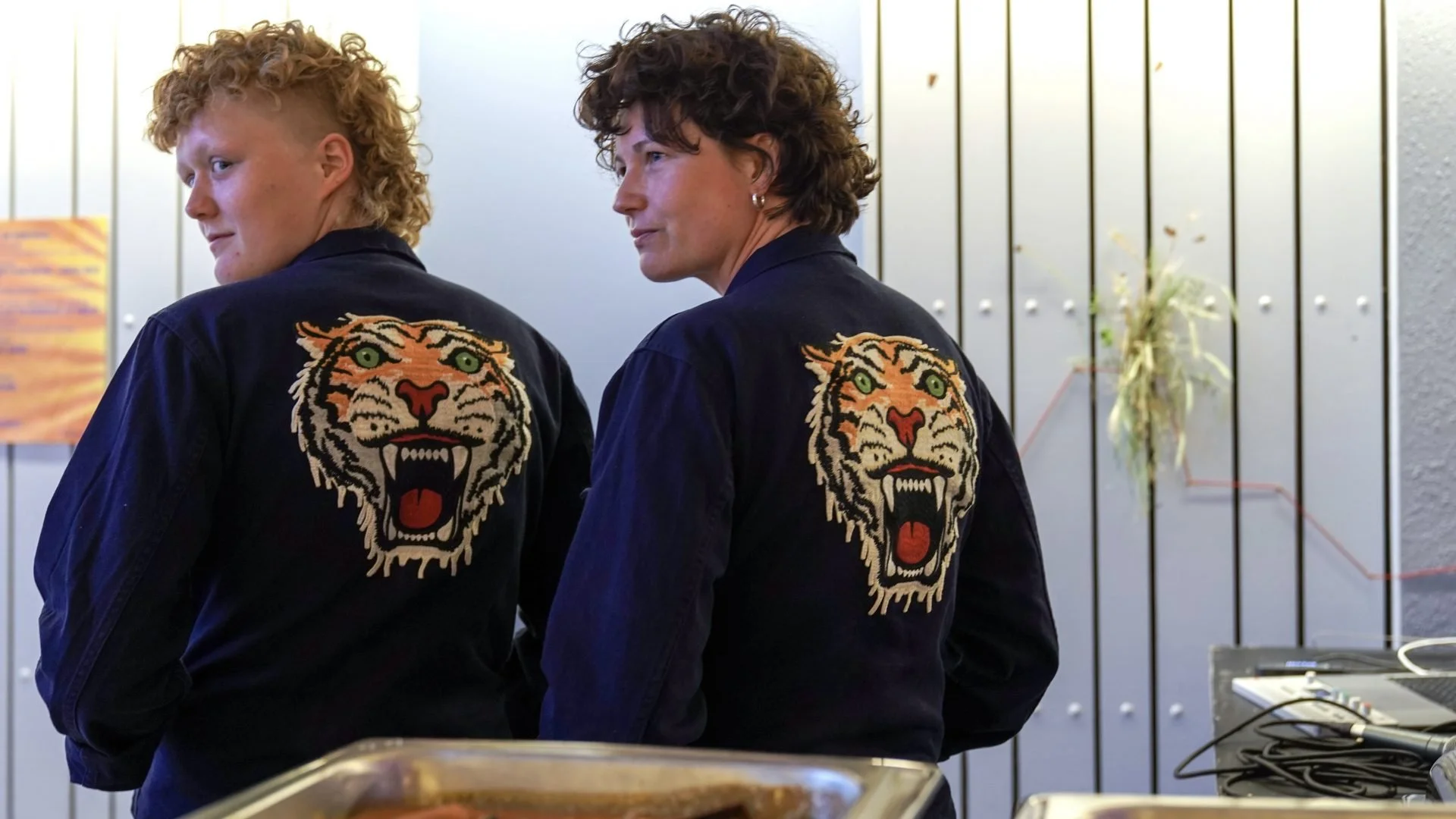 Two women wearing black jackets with a tiger face on the back, standing side by side, in an indoor setting with metal wall panels and some plants in the background.