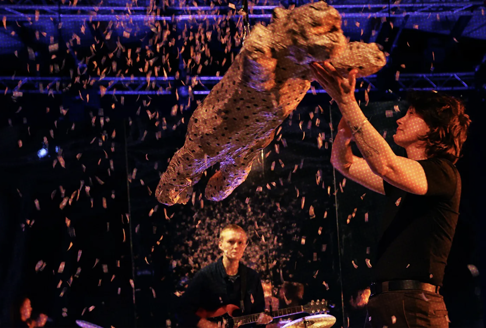 A performer flying through the air on stage with confetti falling, while a woman in a black shirt lifts her with a supporting hand, and a man in the background plays a guitar amid colorful lighting.