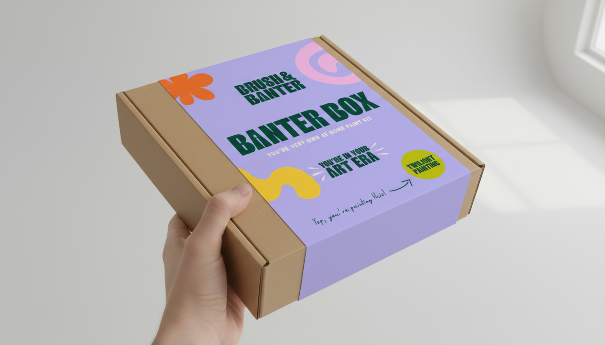 A hand holding a cardboard box with colorful art-themed packaging that reads 'Brush & Bainter, Bainter Box, You're very own at home paint kit, You're in your art era, Transparent painting, Yes, you're painting this!' against a white background and a window in the corner.