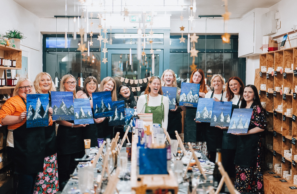 Group of women at a painting class, holding up finished landscape paintings of mountains and lakes, in a bright, decorated studio.
