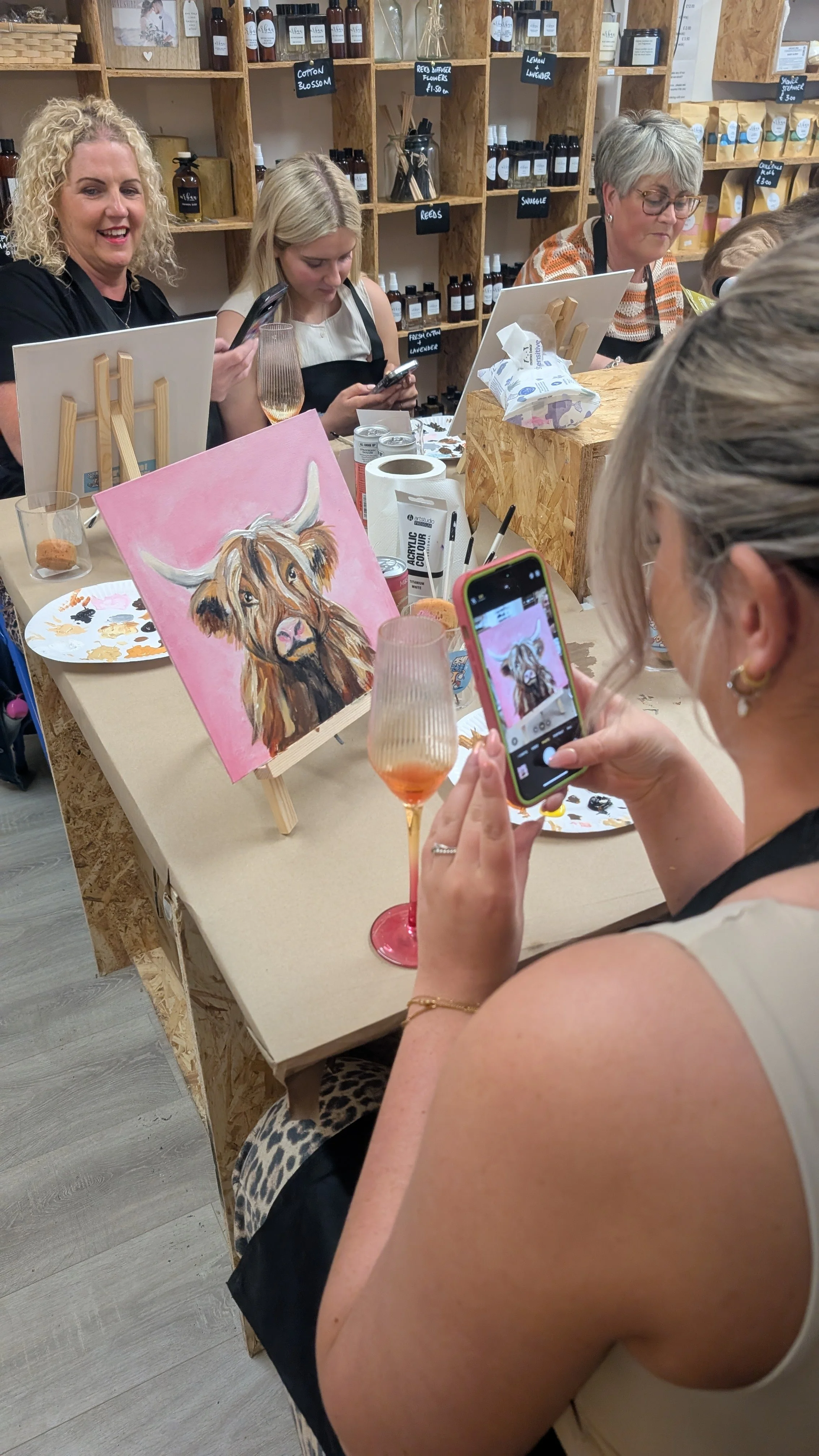 A group of women participating in a paint and sip class, with a painting of a highland cow on an easel, holding glasses of wine, and taking photos, in a cozy craft studio with shelves of bottles and supplies.