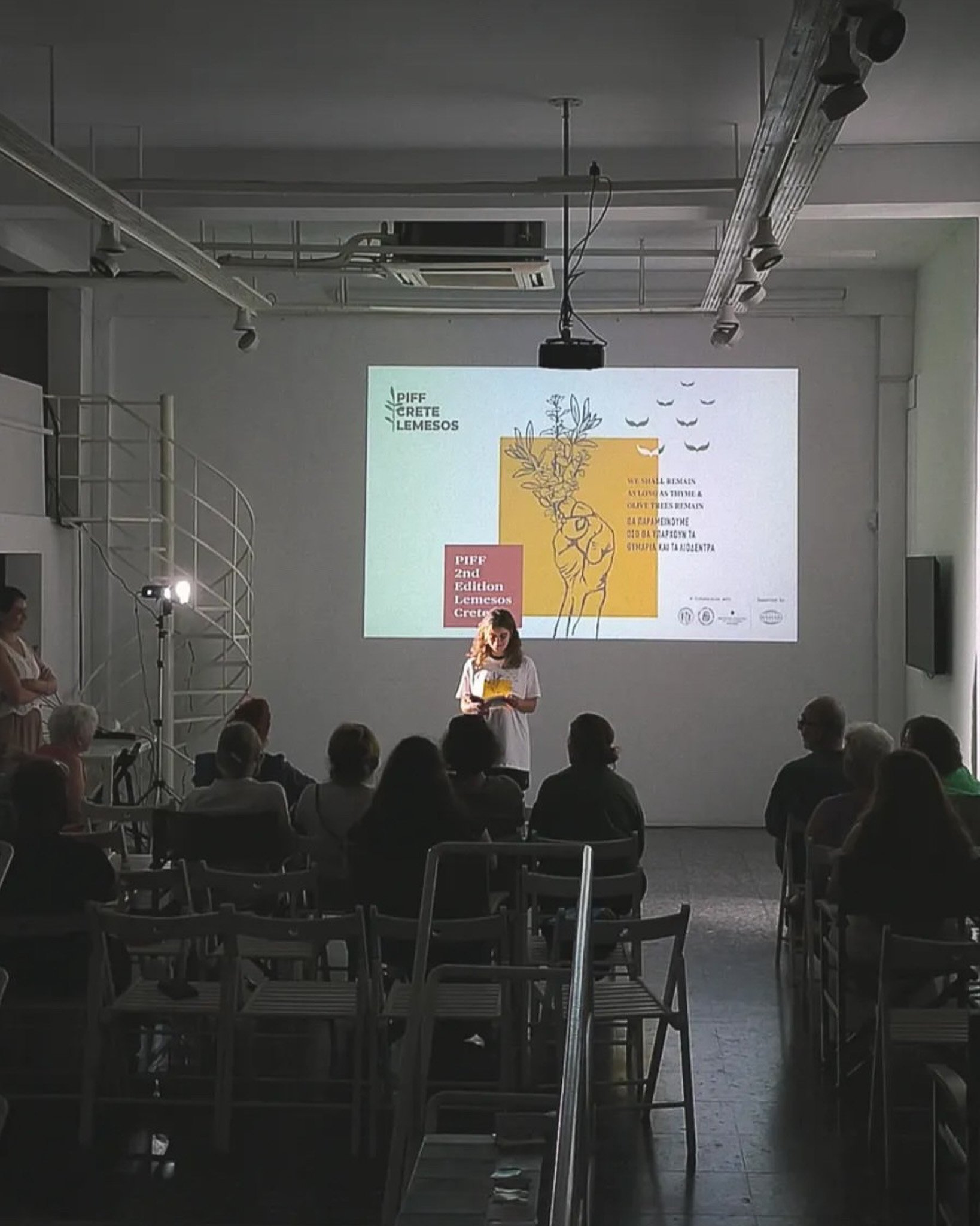 	
PIFF 2nd Edition Limassol Cyprus. Poetry reading at NeMe Arts Centre. A woman is standing in front of a projection screen giving a presentation in a conference room. The projection displays logos and text related to the Lemesos Cultural Center.