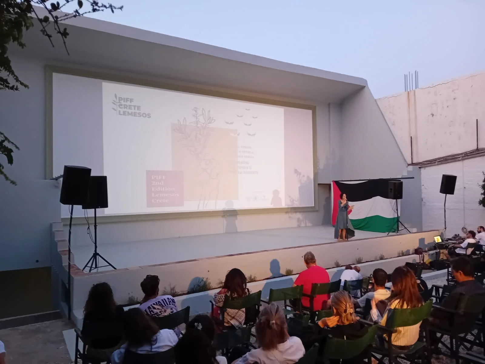 PIFF 2nd Edition. An outdoor movie screening event  A woman is speaking or presenting on the stage. The screen displays a presentation with the logo and text related to the event, Piff Crete Lemesos, and a drawing of a tree and flying birds.