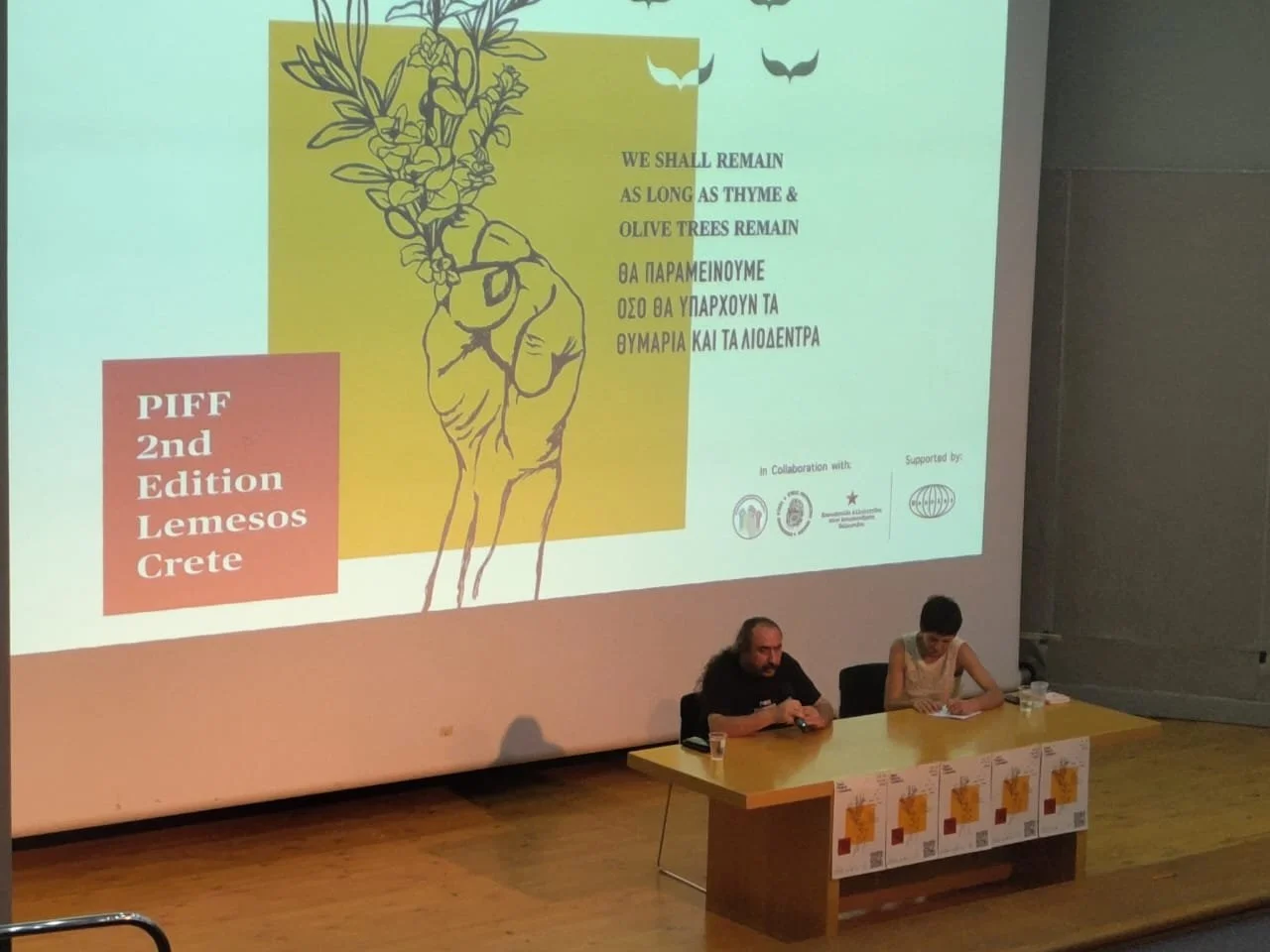 PIFF 2nd Edition Chania Crete. A presentation at a conference with a large screen displaying text and graphics, and two people seated at a table in front of the screen.