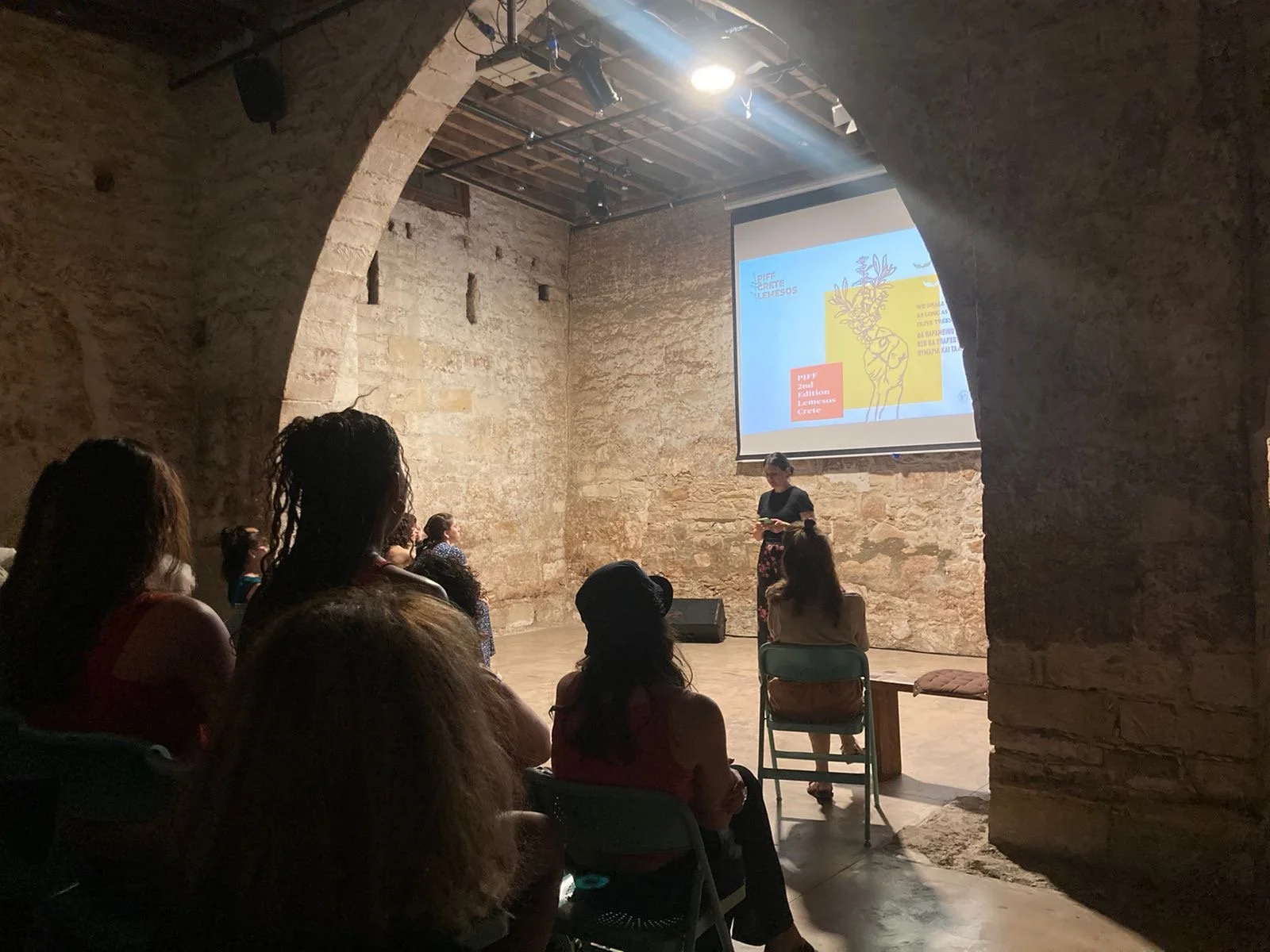Palestinian independent film festival PIFF 2nd Edition Opening night Limassol Theatre. A speaker and a woman are standing in front of an audience in a theater or lecture hall. The audience is seated on chairs and tiered seats, attentively listening.