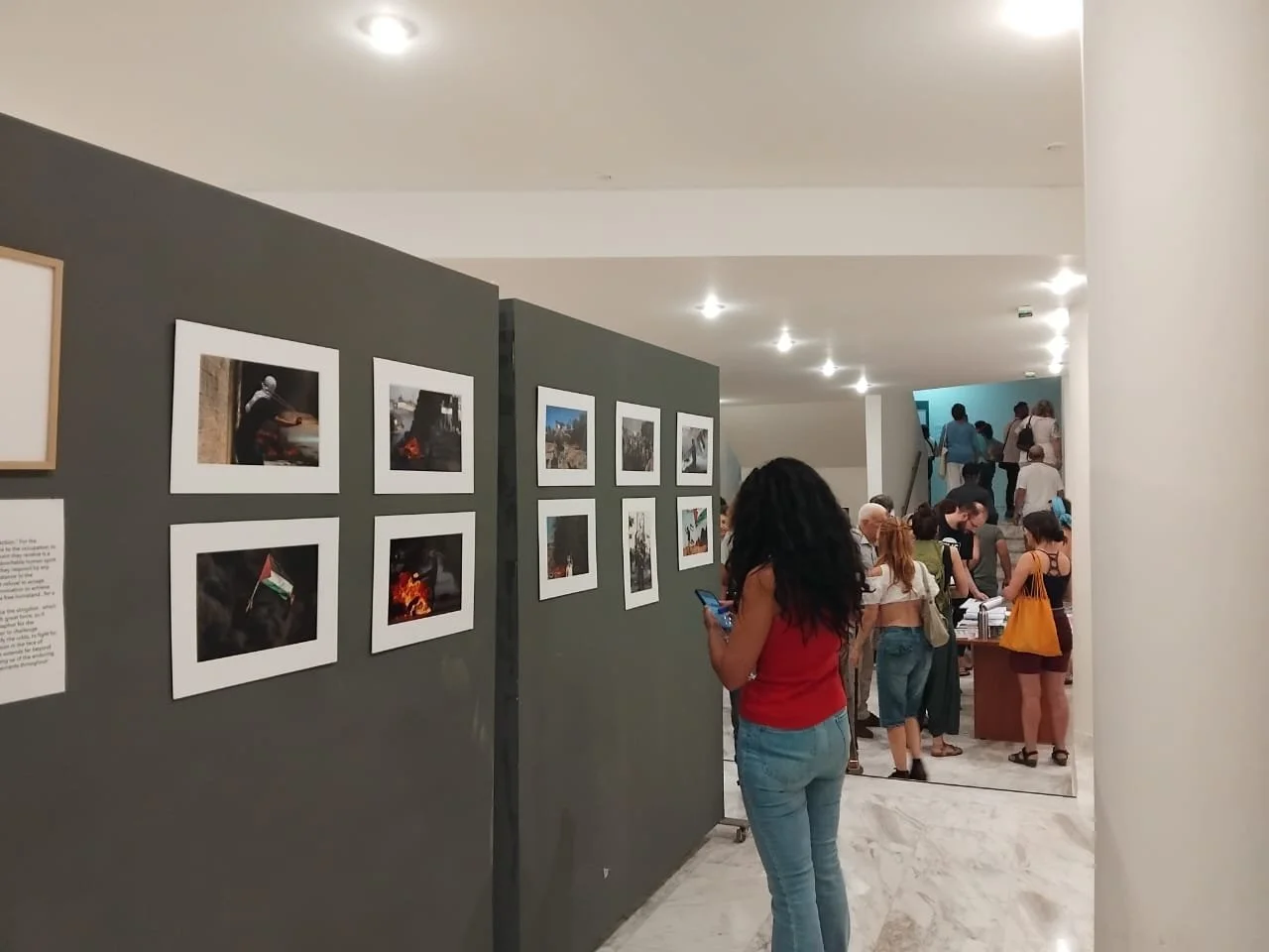 	
PIFF 2nd Edition Chania Crete. Photography Exhibition. People viewing photographs and artwork on display in an art gallery, with a woman in a red top and jeans looking at her phone.