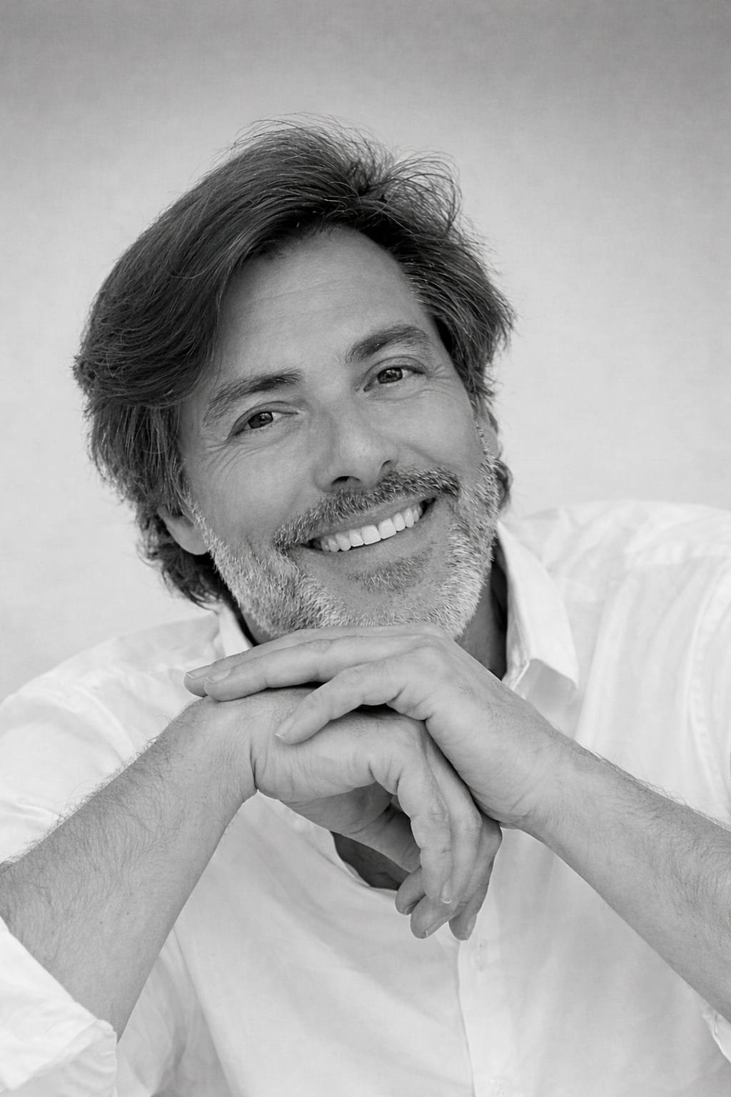 Black and white portrait of a smiling middle-aged man with wavy hair and a beard, resting his chin on his interlaced hands, wearing a white shirt.