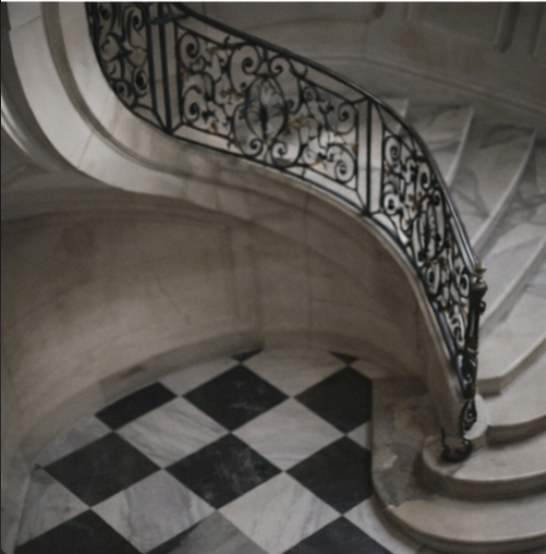 A curved marble staircase with an ornate black metal railing, overlooking a black and white checkered tile floor.