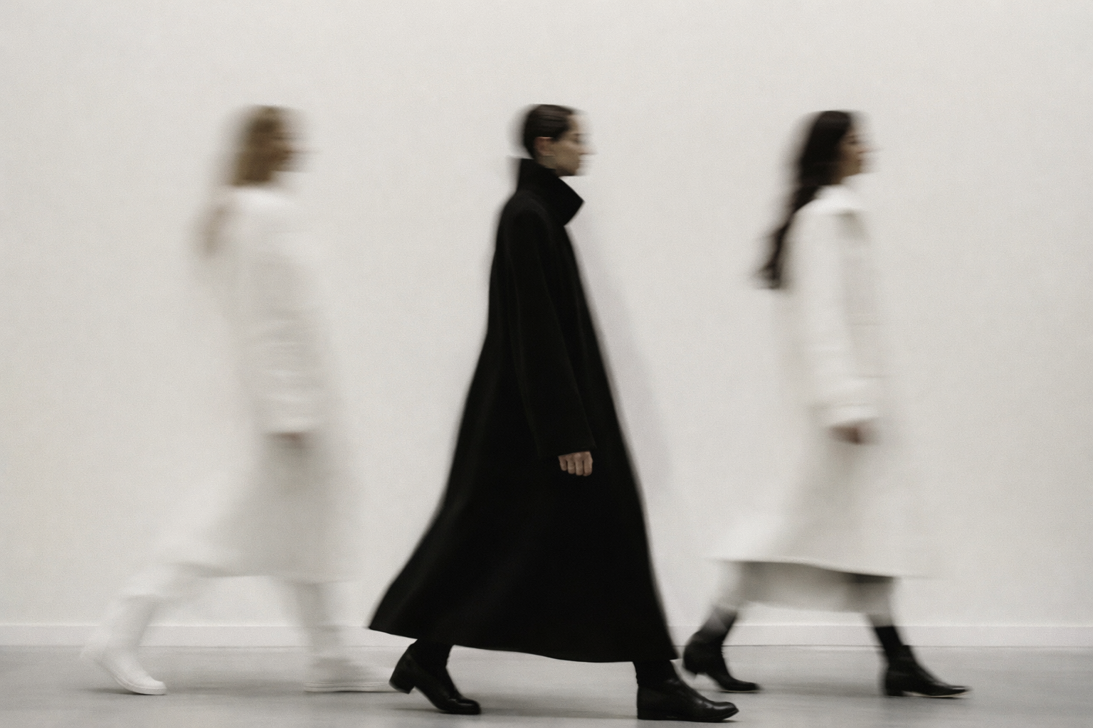 Three people walking in a line, all dressed in black and white, with the person in the middle wearing a long black coat.