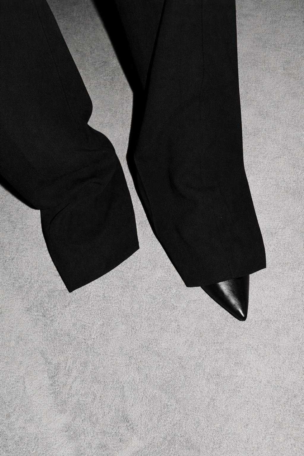 Part of a person's black suit and black pointed-toe shoe, standing on a grey carpeted floor.
