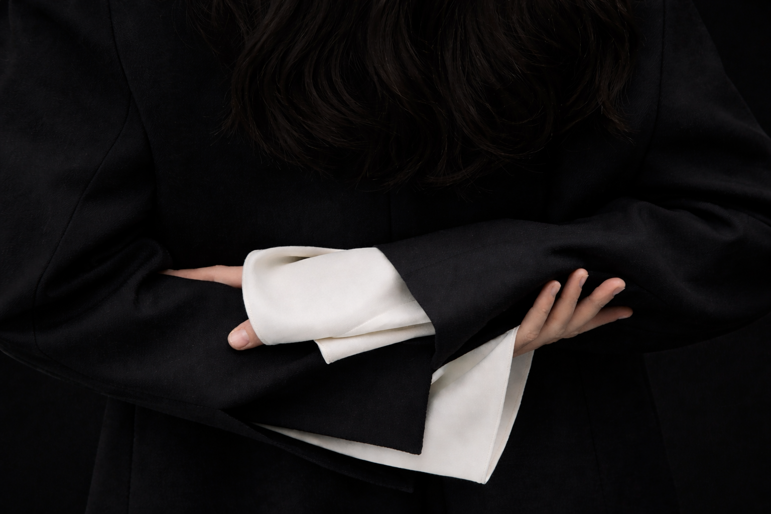 Close-up of a woman with dark hair, dressed in black, with her arms crossed. She is wearing a white shirt under the black jacket.