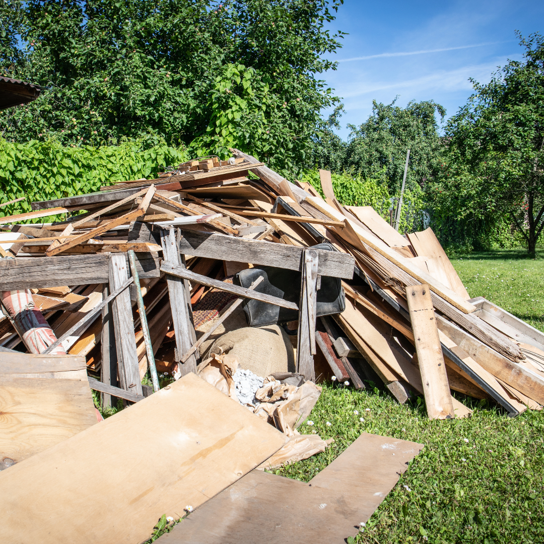 Pile of discarded wood and debris outdoors on grass, with trees and a clear blue sky in the background.