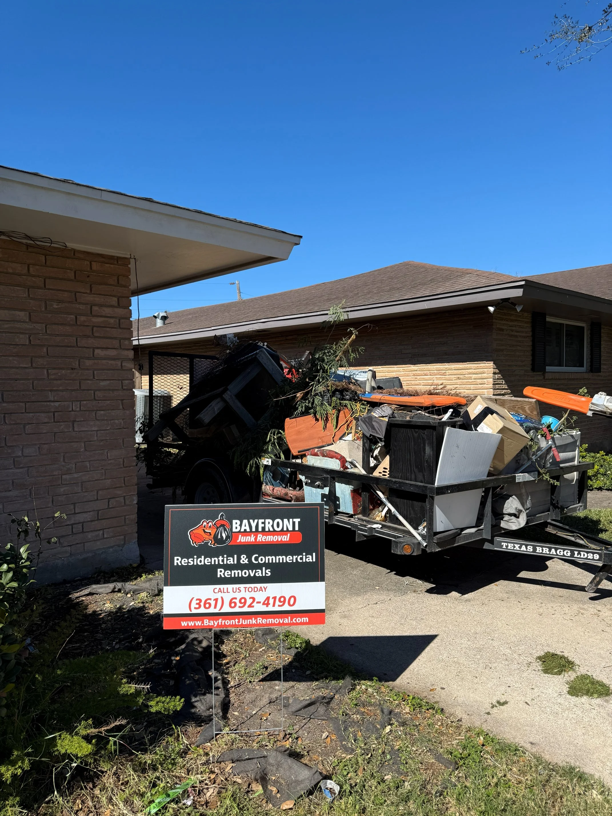 A trailer filled with household debris and junk parked in front of a brick house, with a sign advertising residential and commercial junk removal services from Bayfront Junk Removal.