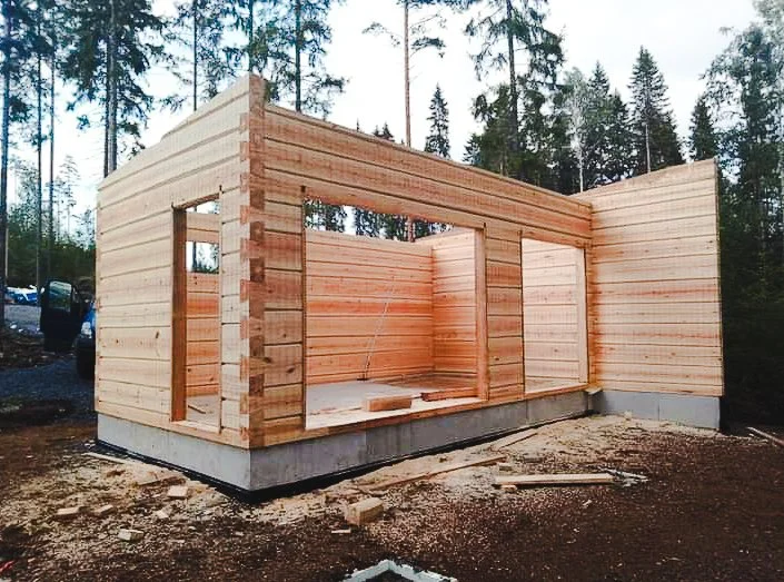Wooden house under construction with exposed frame and walls, situated on a concrete foundation in a wooded area.
