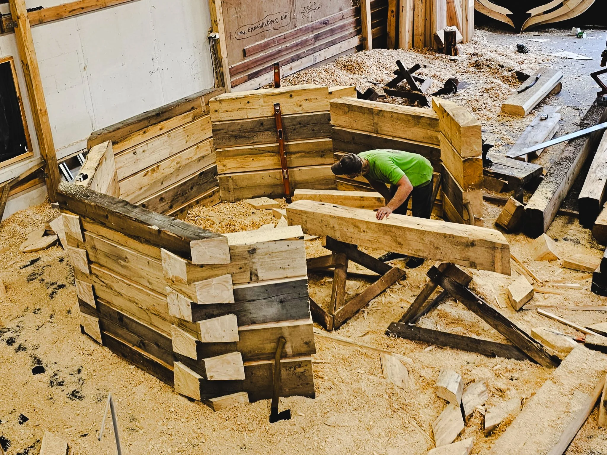 A person in a green shirt is working on building a circular wooden structure indoors, surrounded by wood, sawdust, and construction tools.