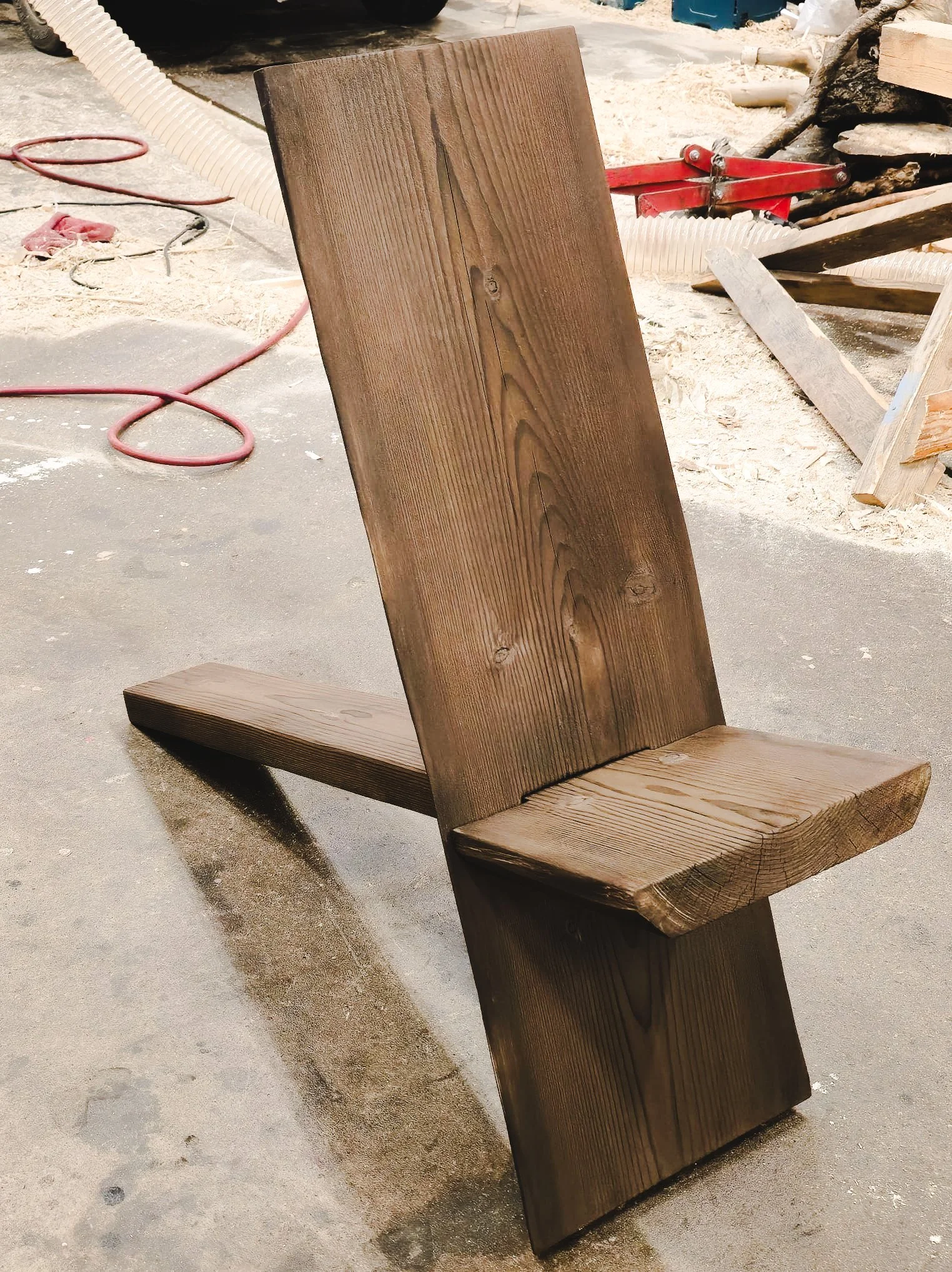 A wooden bench with a backrest, made from two large stained wood planks, situated on a concrete floor in a woodworking workshop or garage. In the background, there are woodworking tools, sawdust, and a hose.