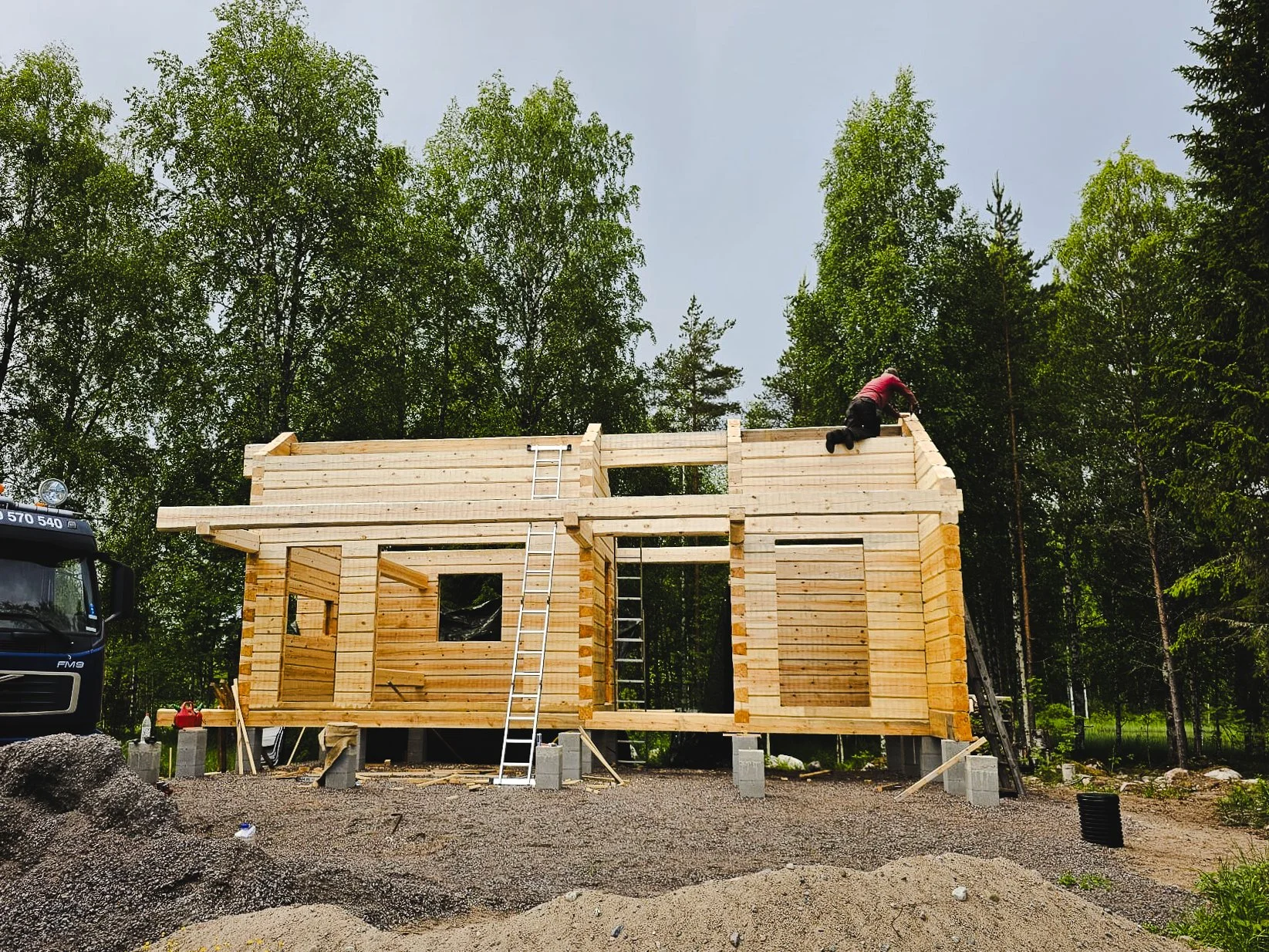A wooden house under construction with two workers on the roof, surrounded by trees and construction materials.