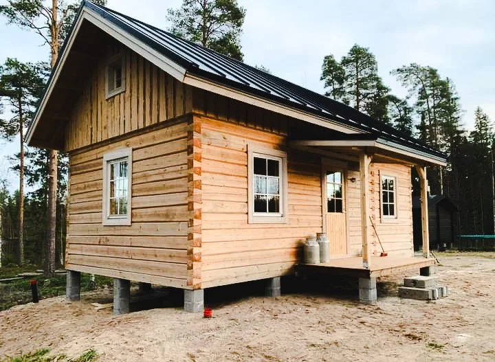 Unfinished wooden house on stilts with a black metal roof, surrounded by trees, during daylight.