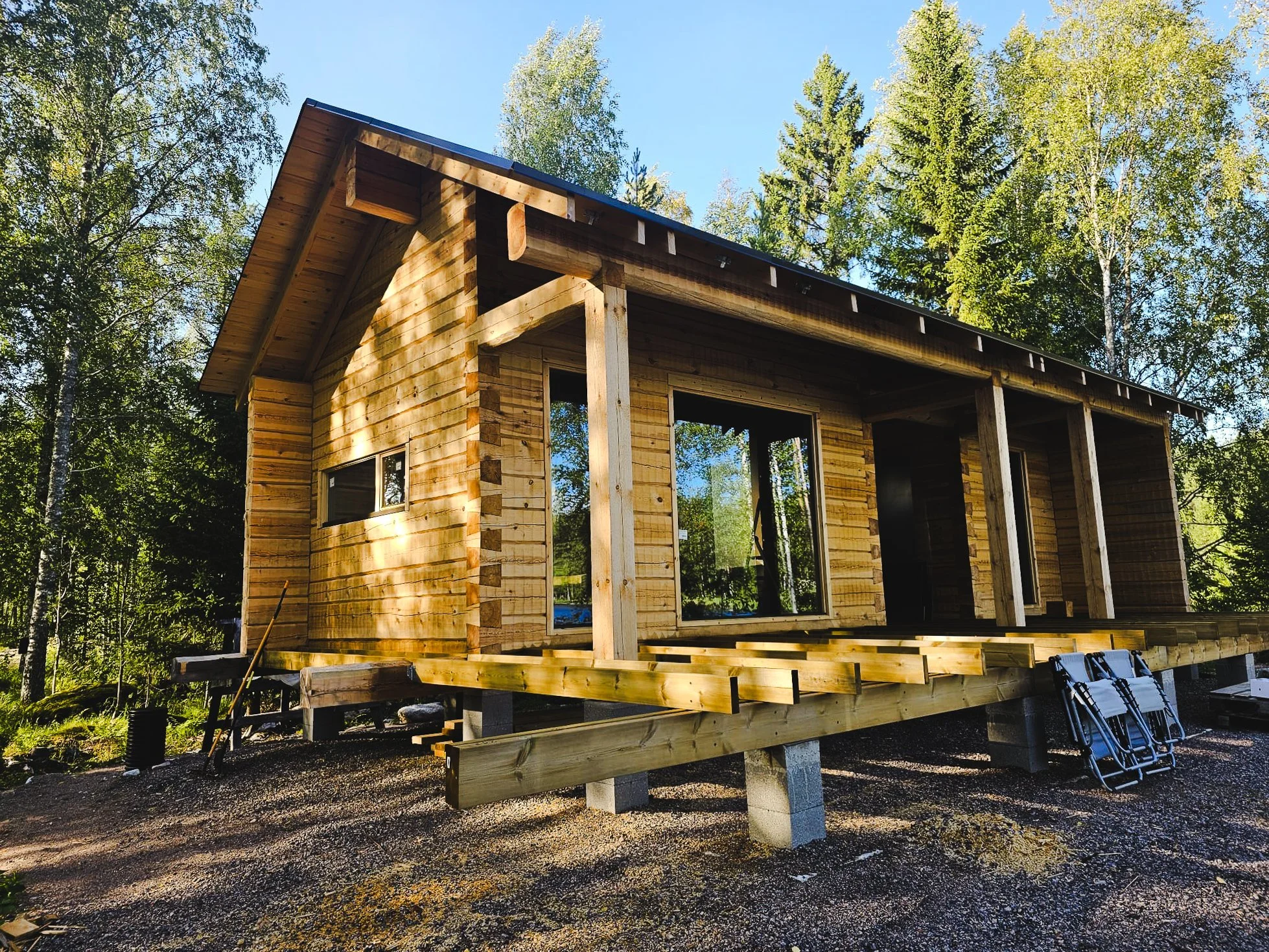 A wooden house under construction in a forested area with trees and blue sky visible in the background.