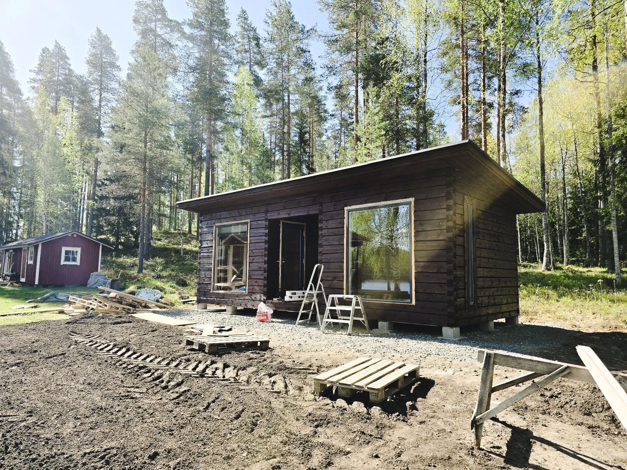 Construction site with a small dark wooden house in a forested area, surrounded by trees, with construction materials and tools nearby.