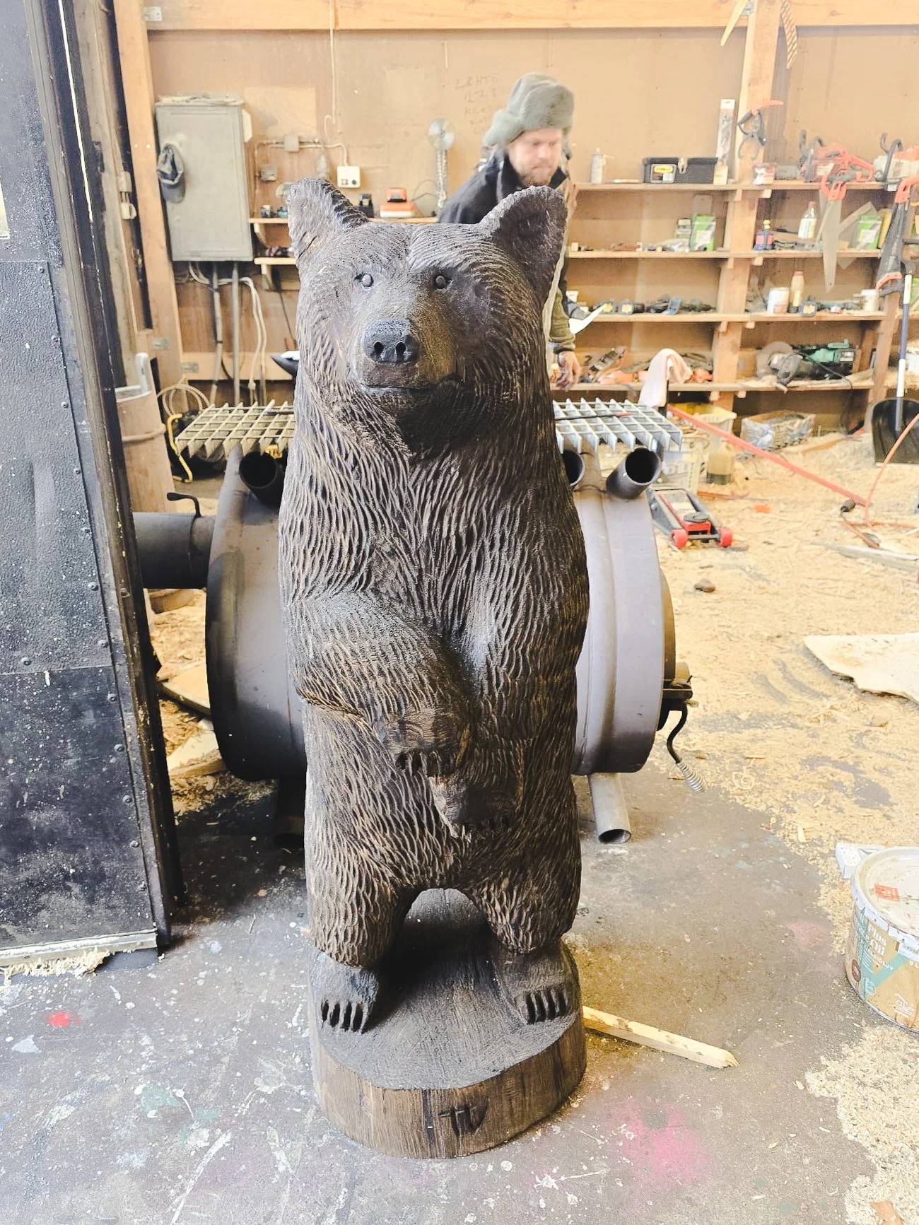 Wooden bear sculpture in a workshop, with a man working in the background surrounded by tools and woodworking supplies.