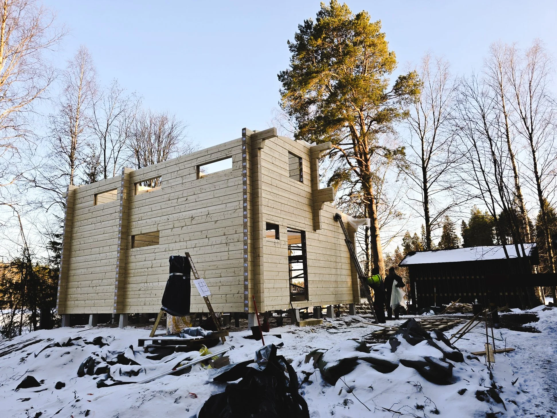 Wooden house under construction in a snowy outdoor setting with trees in the background and workers on site.