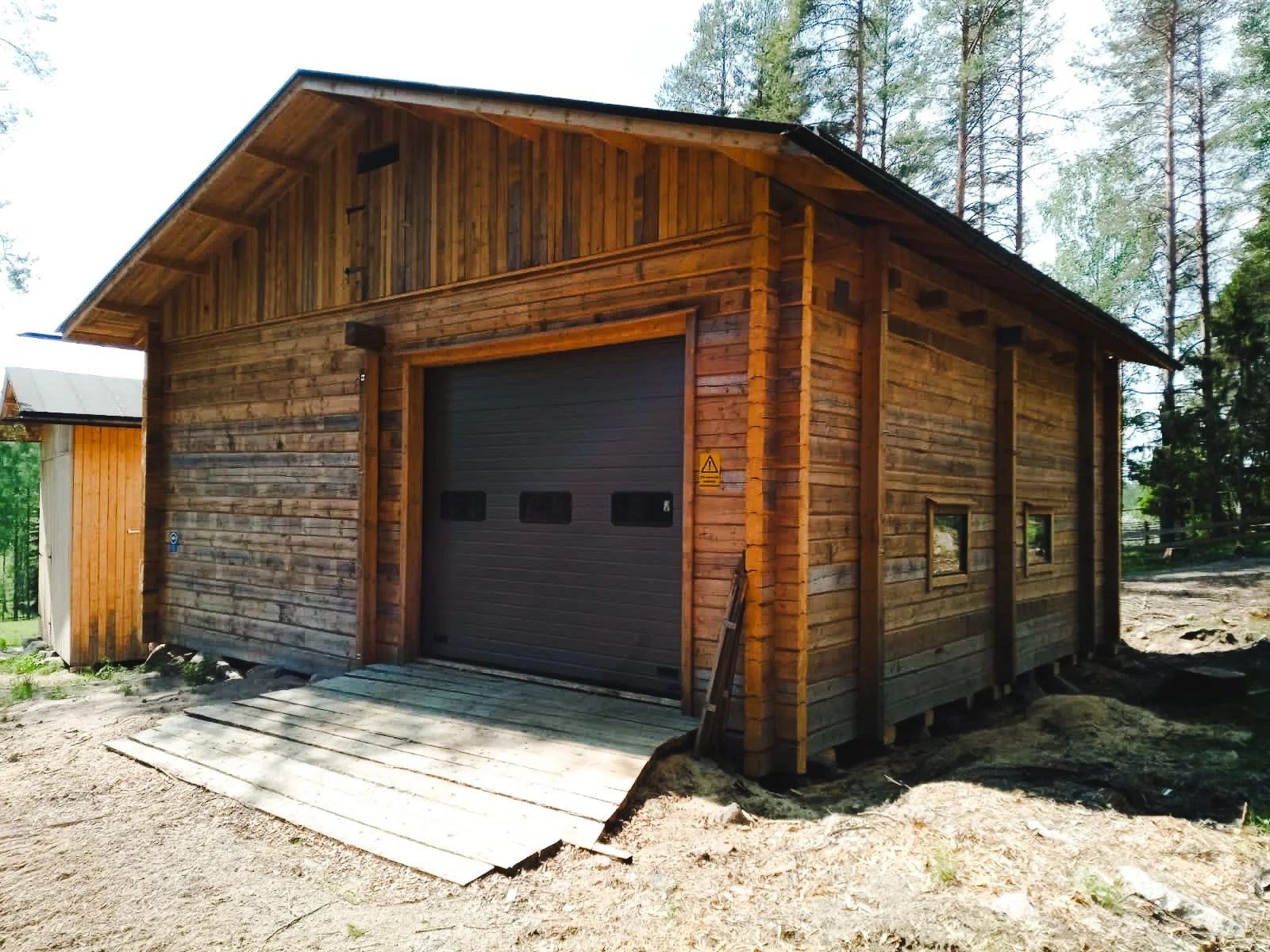 A wooden garage with a black roll-up door, surrounded by a wooded area with trees and a dirt ground.