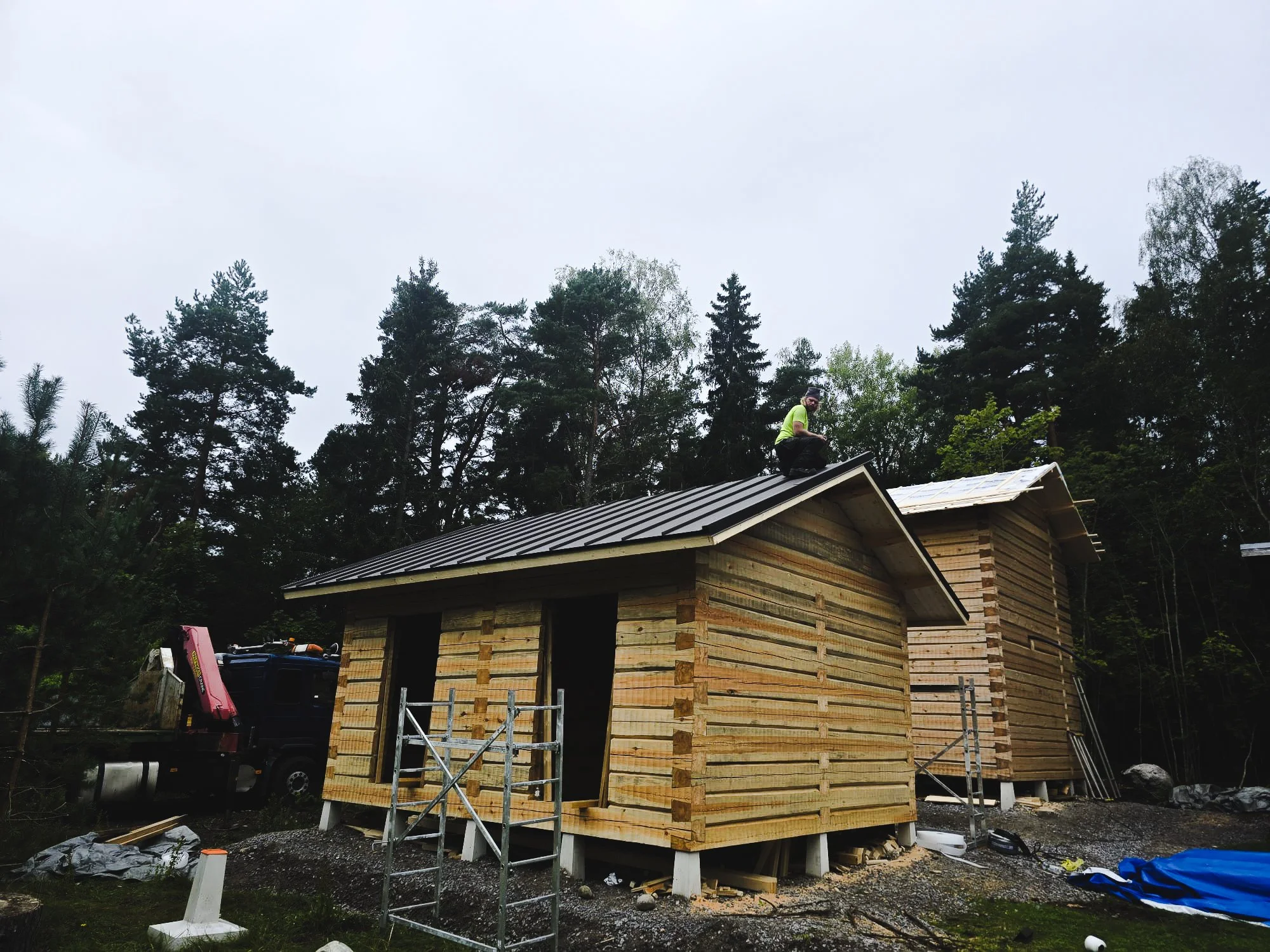 Two small wooden houses under construction in a forest, with a worker on the roof of the front house, and construction materials and a truck nearby.