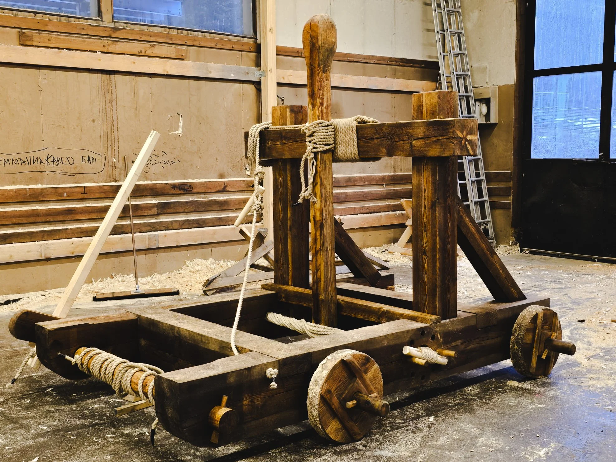 A large wooden medieval siege weapon, possibly a ballista or catapult, built from heavy timber with large wheels and ropes, inside a workshop or warehouse.