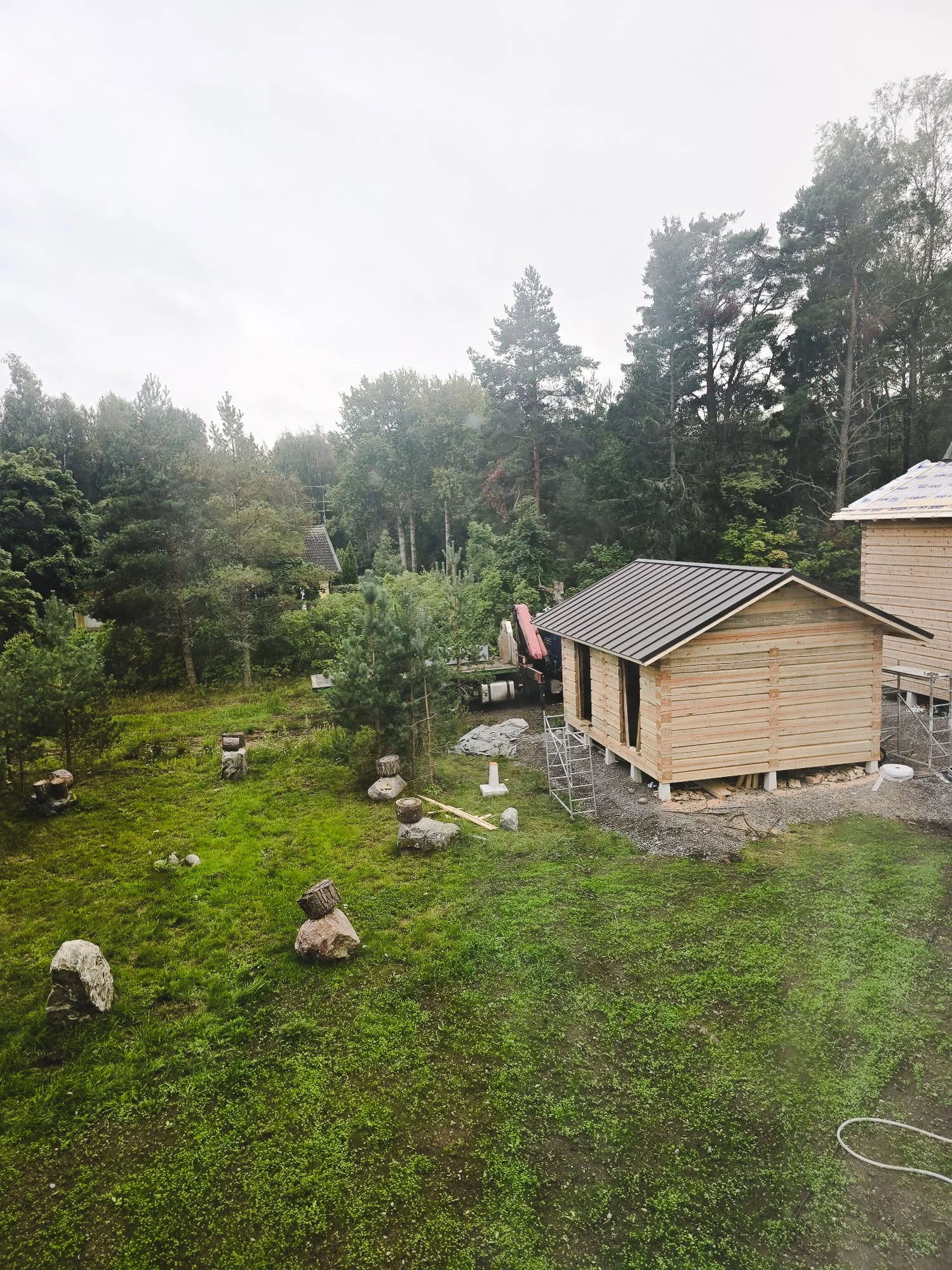 Construction site with a wooden shed being built, surrounded by trees and grass, on a cloudy day.