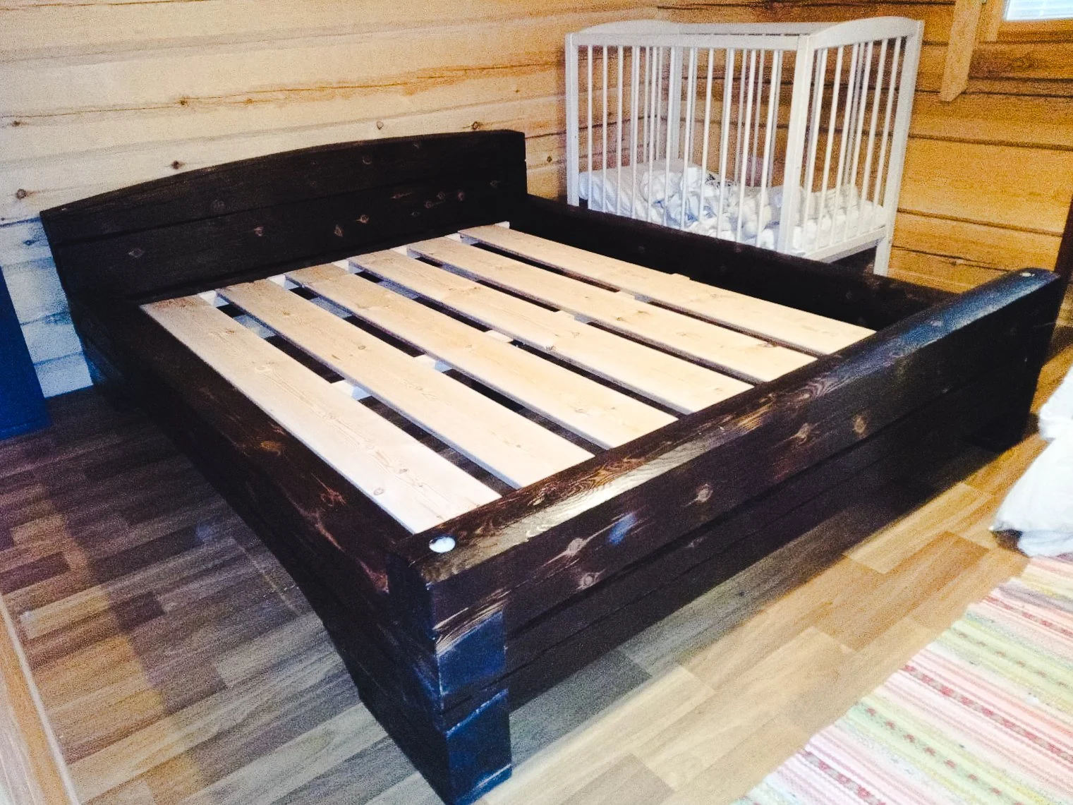A wooden bed frame with a dark finish in a rustic room with wood-paneled walls, next to a small crib with a white railing and bedding.
