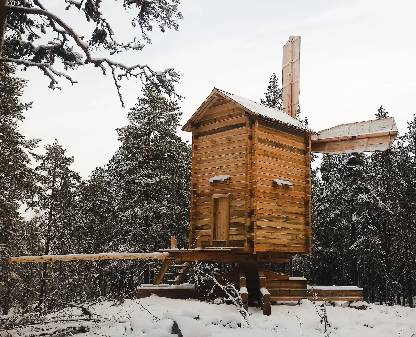 A small wooden windmill structure in a snowy forest with snow on the ground and trees.
