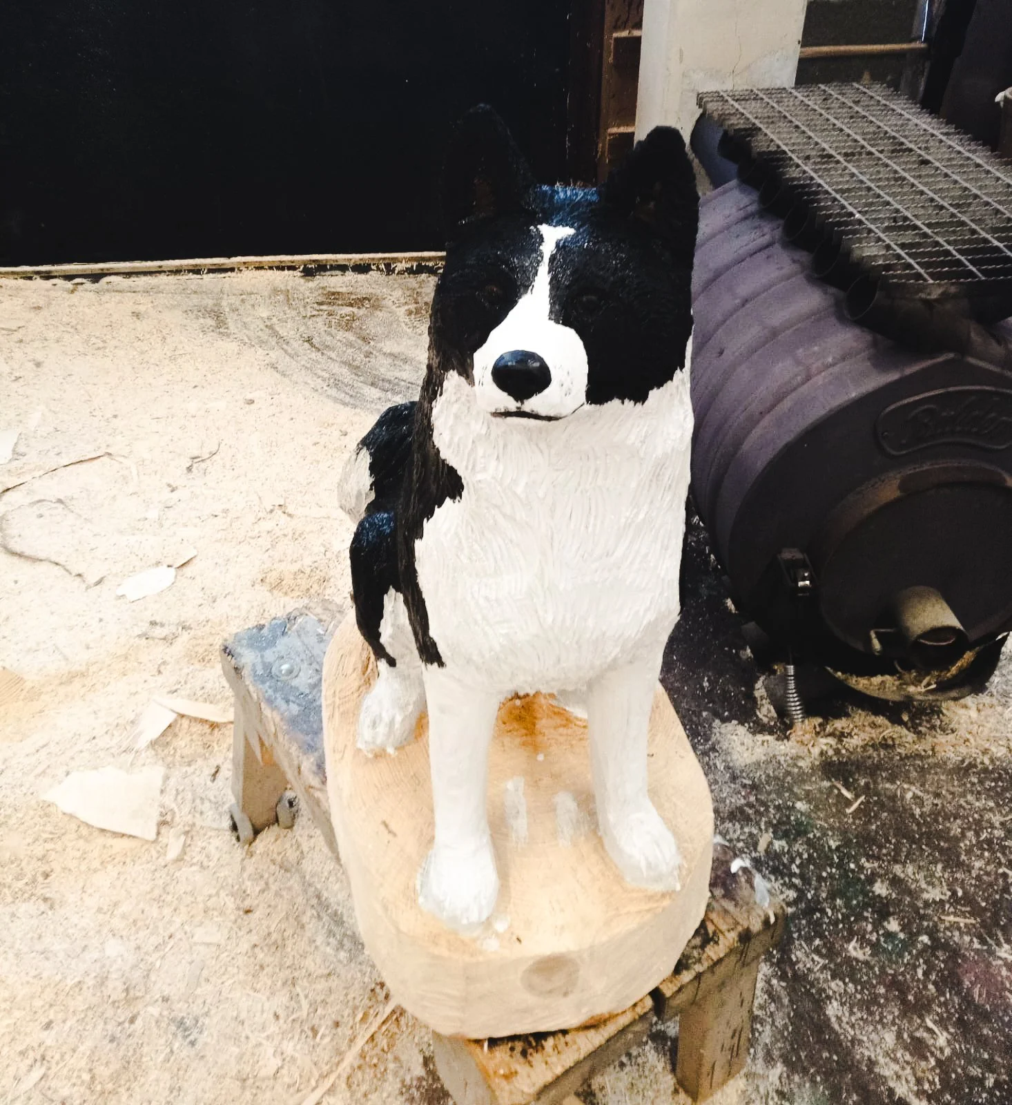 A carved wooden sculpture of a black and white Border Collie dog on a wooden stand, located in a workshop with sawdust on the floor and a large pipe or vent in the background.