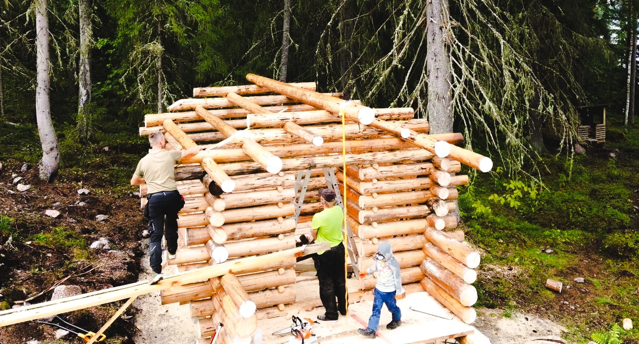 People building a log cabin in a forest, with trees and greenery surrounding the construction site.