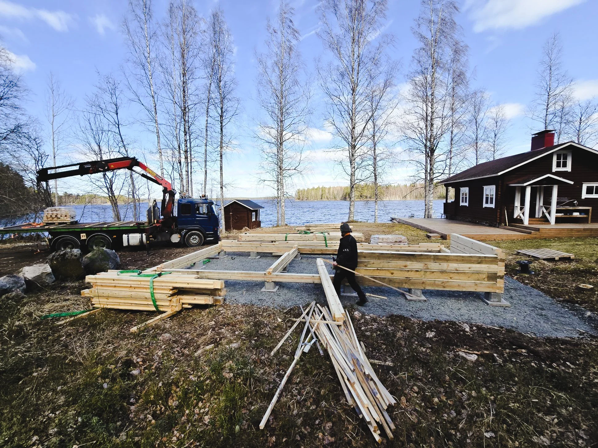 Construction site near a lake with wooden framing, a worker, a house, and trees.