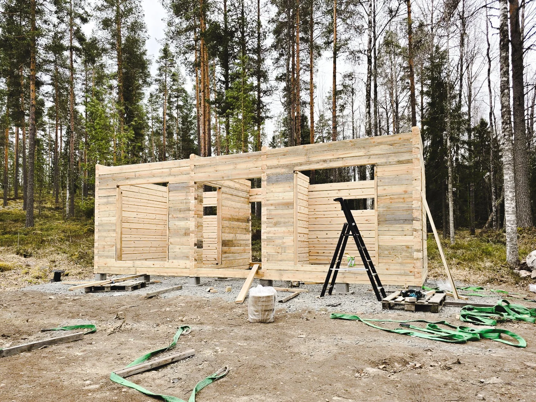 Unfinished wooden house frame under construction in a wooded area, with tools and construction materials around.