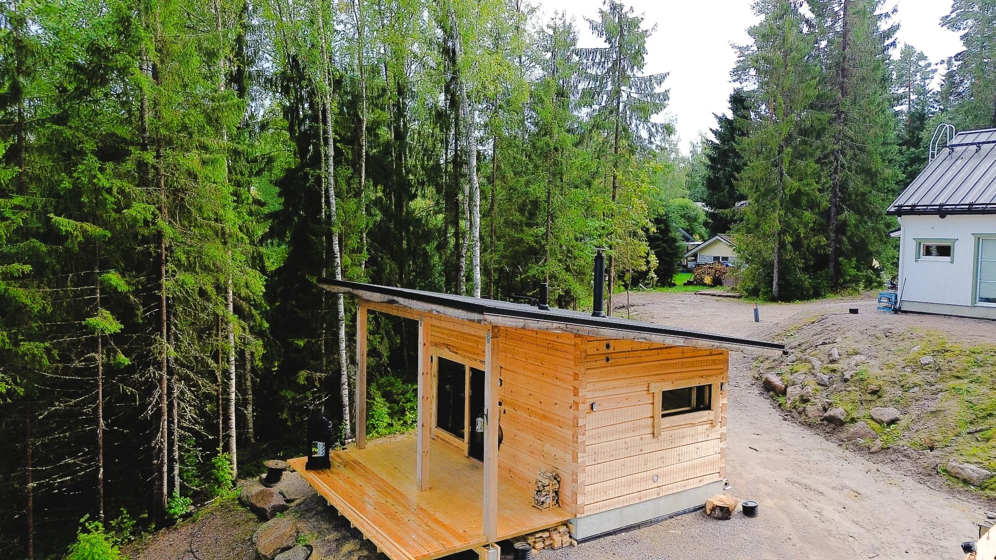 A small wooden cabin under construction in a forested area with tall green trees, with a neighboring house visible in the background, and a dirt pathway leading to the cabin.
