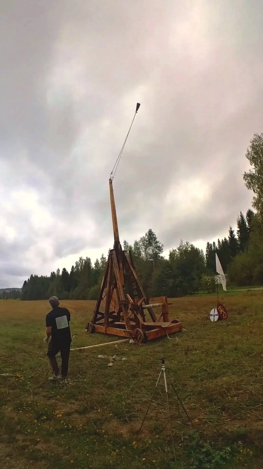 A person in a black shirt and gray cap is on a field, operating a wooden catapult with a long arm and wheels.