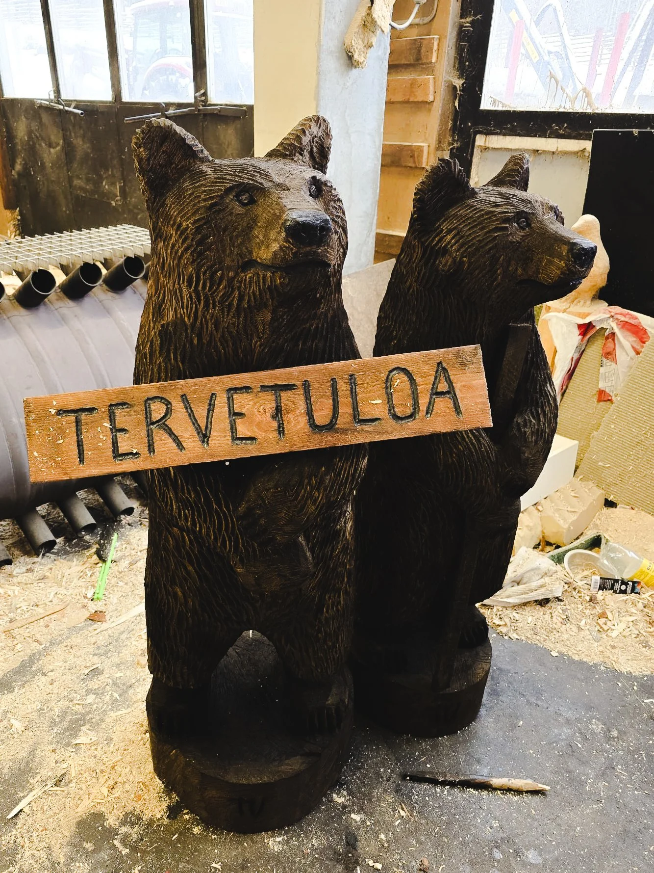 Two carved wooden bear sculptures holding a sign that says 'TERVETULOA' in Finnish, in an indoor workspace with wood shavings and tools scattered around.