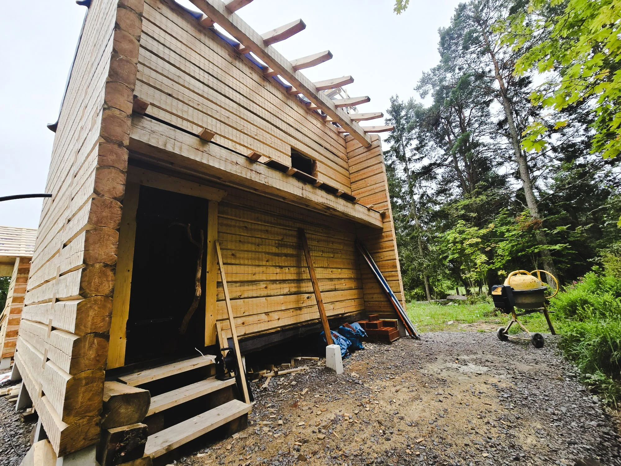 Construction site of a wooden house with unfinished walls and a small staircase outside, surrounded by trees and construction tools, including a cement mixer.