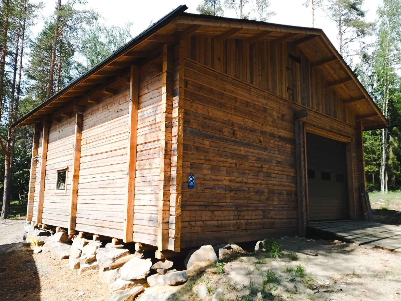 A wooden barn with a sloped roof, built on large rocks, surrounded by trees in a forested area.