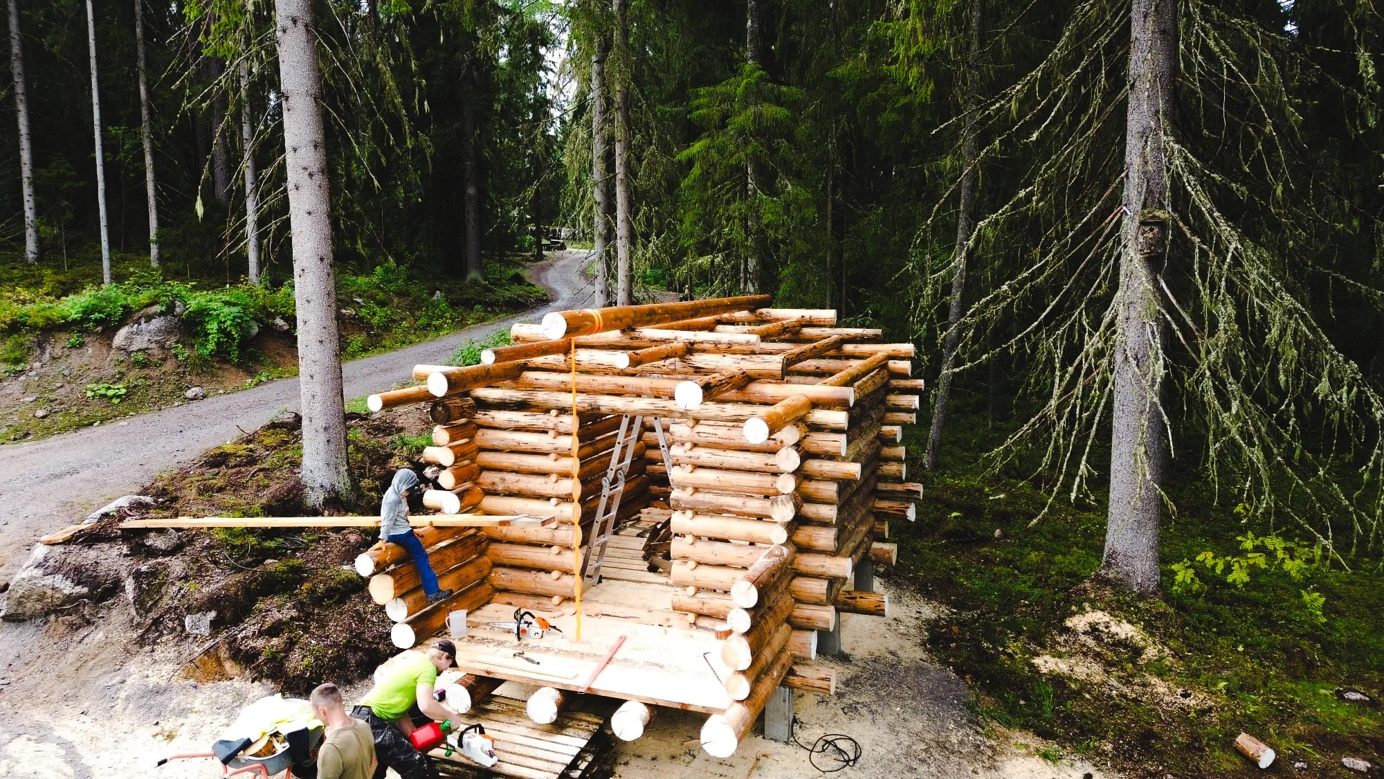 People assembling a log cabin in a forested area with tall trees and a winding dirt road in the background.