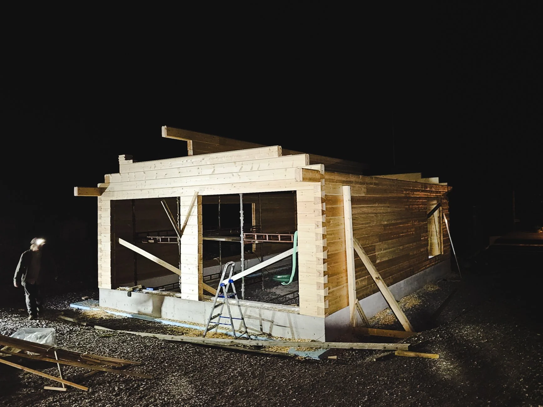 Construction site at night with a partially built wood house, a person standing nearby, and construction tools and materials around.