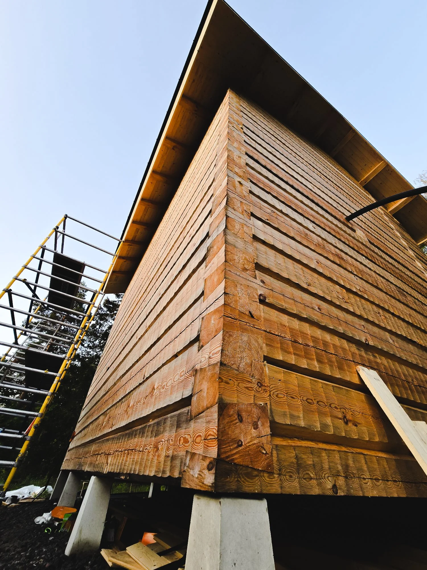 Building under construction with wooden planks and concrete supports, scaffolding on the side, against a clear sky.