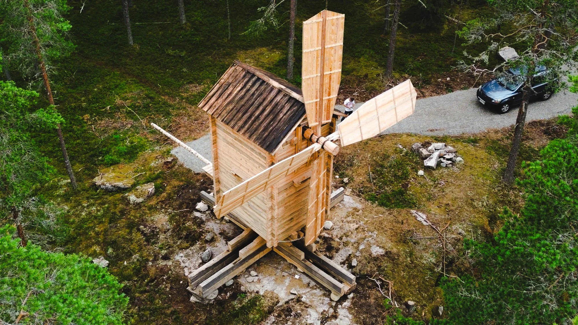 A wooden windmill under construction in a forested area, with a car parked on a gravel path nearby.