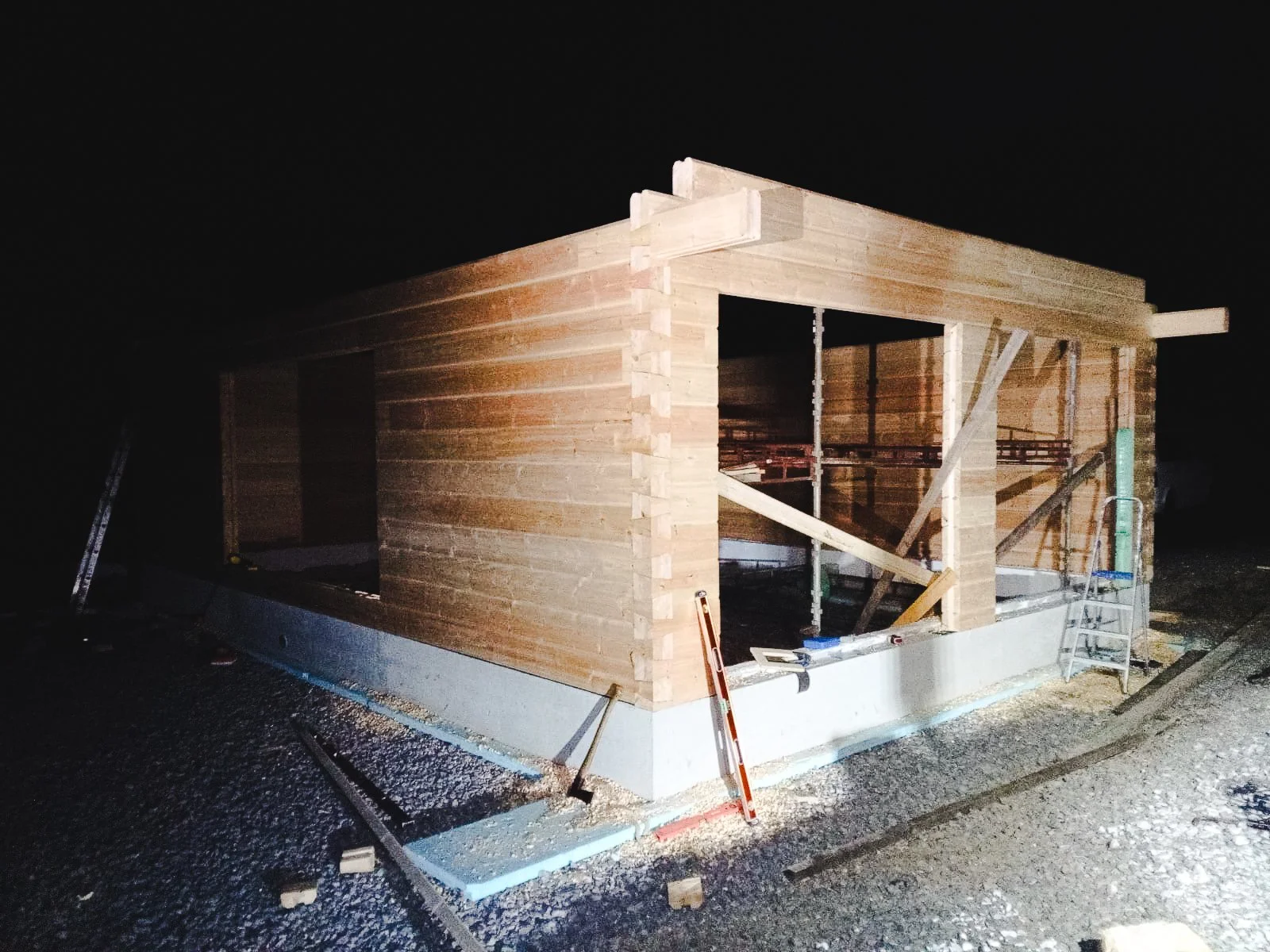 Nighttime photo of a small wooden house under construction with scaffolding, ladders, and construction tools around it.