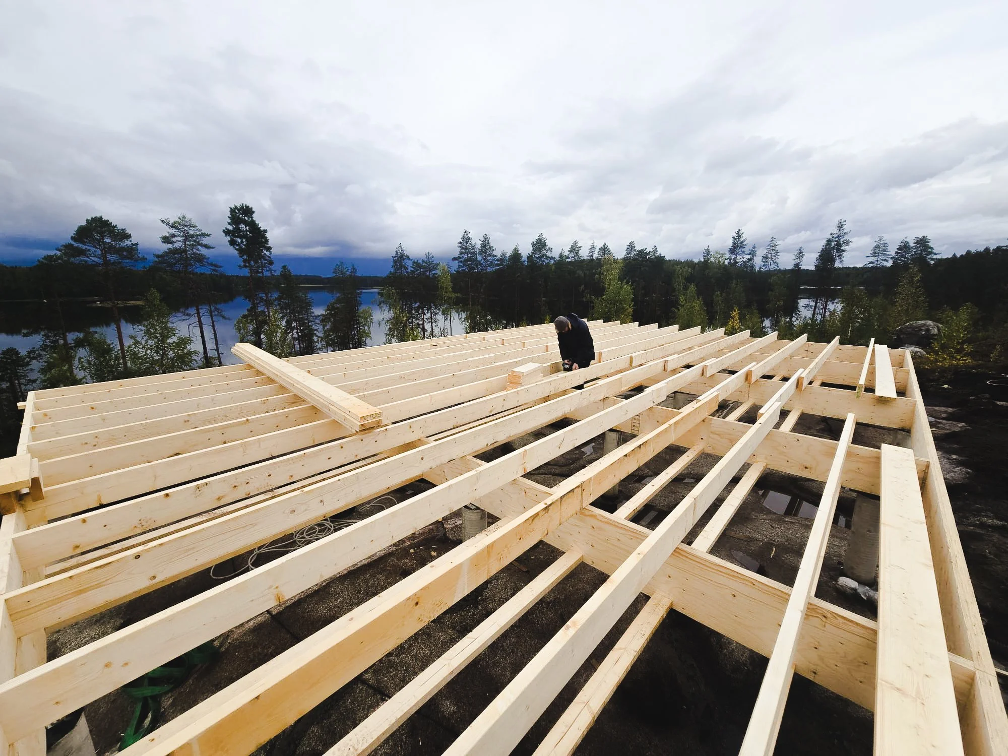 Person working on framing wooden structure outdoors near a lake with trees and cloudy sky.