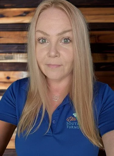 A woman with long blonde hair and green eyes wearing a blue polo shirt with a logo, standing in front of a wooden background.